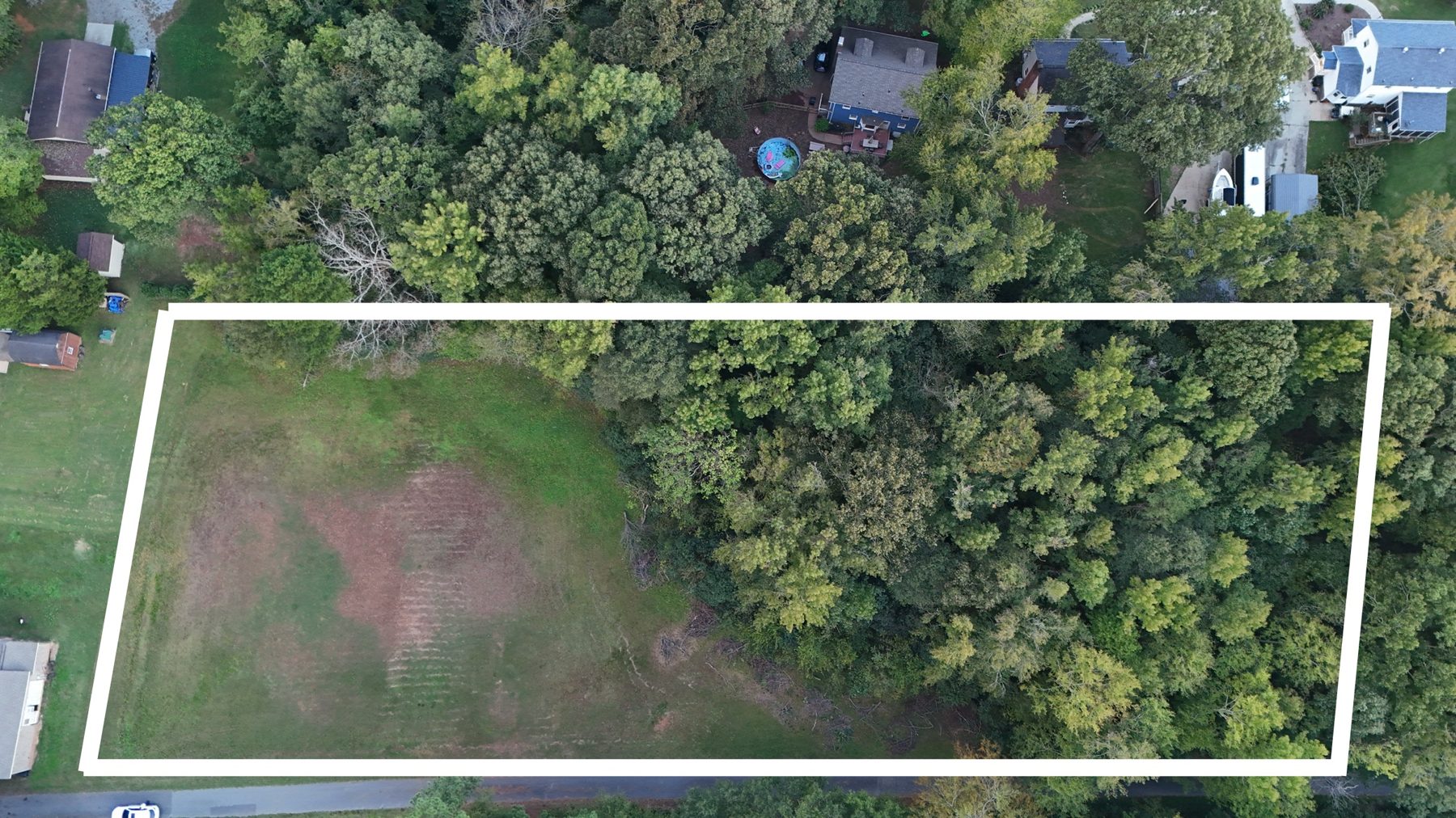 Drone view straight down on the lot, an open field on the left and hardwoods on the right, a paved lane along the bottom, its boundary drawn in white.