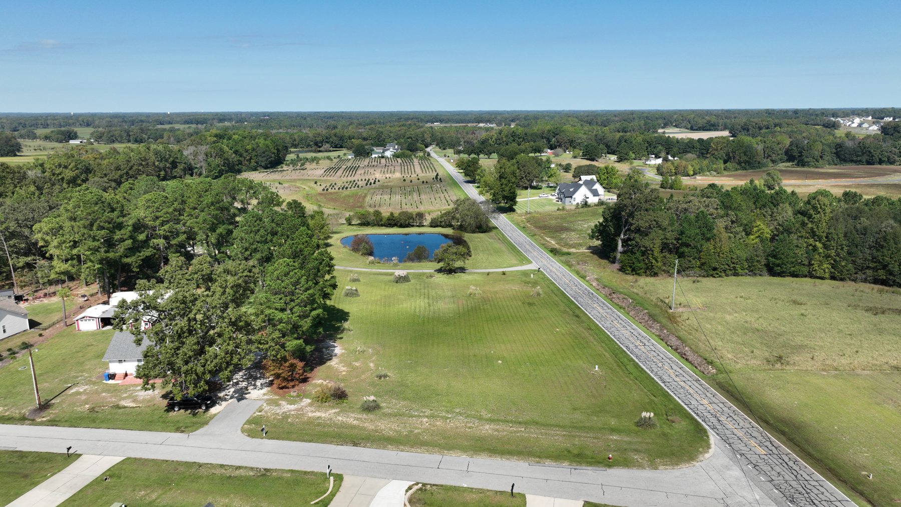 Drone view up Old Stage Road over the mowed corner lots, a pond and farm fields beyond.